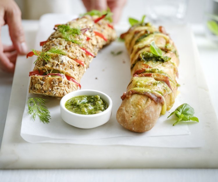 Broodjes op de BBQ Twee broodjes liggen op een snijplank. Je ziet dat het twee versies zijn, nl een broodje met groene pesto, sneetjes ham en kaas. En dan nog een versie met roomkaas, paprika en rode ui. Extra pesto staat in een potje bij de broodjes, voor wie wat extra wil. Een vrouw zet de plateau of snijplank op tafel, haar handen zijn nog in beeld.