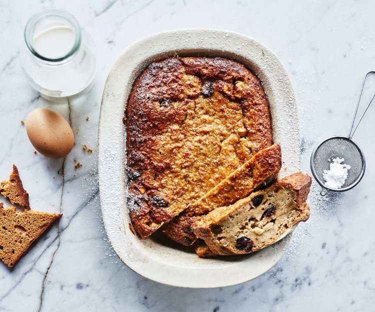 Een heerlijke homemade broodpudding staat op een marmer aanrecht in een witte ovenschaal. Hij is al aangesneden en een stukje uit de vorm genomen. Om de foto meer boddy te geven ligt er een eitje, flesje melk, stukje peperkoek en bloemsuiker naast.