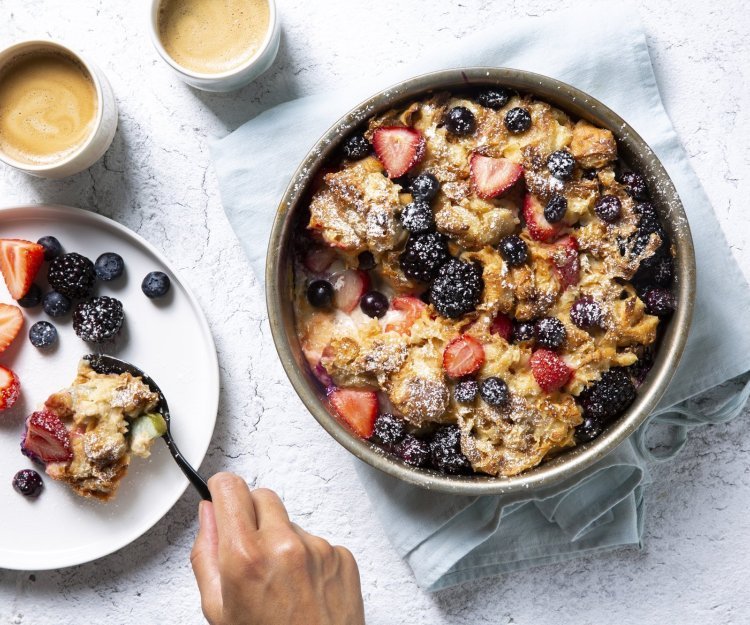 Broodpudding met rabarber, aardbeien, bramen en blauwe bessen Homemade broodpudding met rabarber en zomerbessen staat op tafel. Een vrouwelijke gast schept haar een portie uit om volop te genieten van dit dessert met een kopje koffie.