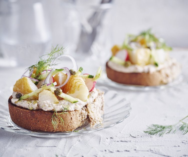 Een goedgevulde bruschetta ligt op een klein glazen bordje en staat op tafel. De bruschetta werd afgewerkt met een takje dille. Achteraan de tafel staat een kan met water en bestek om deze toast te verorberen. 