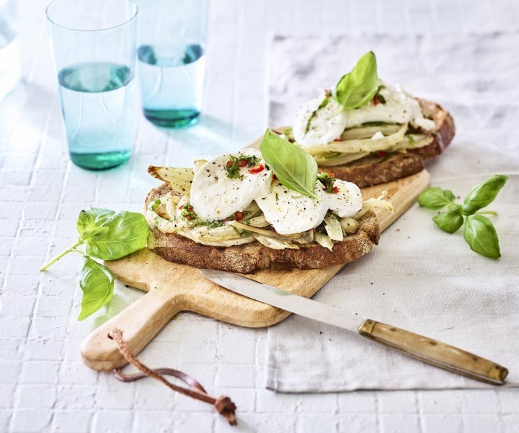 Bruschetta met geroosterde venkel en mozzarella Een rijkbelegde toast met venkel en mozzarella liggen op een houten plank. Boven deze plank staan twee blauwe glazen gevuld met water. Het gerechtje is afgewerkt met enkele basilicumblaadjes.