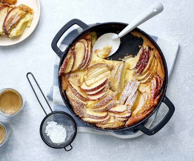 Een clafoutis van appelstukkjes staat op het aanrecht in een zwarte fonte schaal. Er is al een portie uitgeschept en staat op een bordje naar de pot. Er is ook bloemsuiker over het dessert gestrooid en de koffie is al uitgeschonken. 