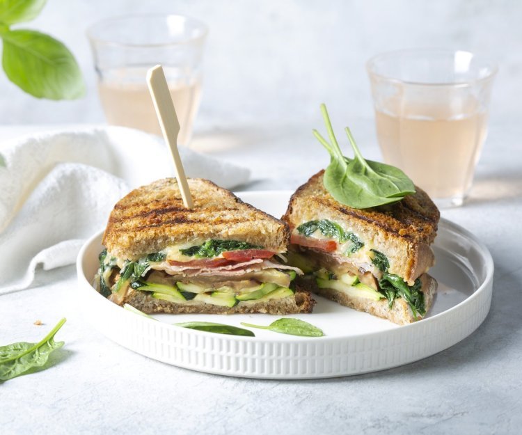 Croque met kaas, spinazie en gegrilde courgette en aubergine Een aangesneden croque ligt op een wit bord. De croque zit boordevol groentjes, en zit vast met een houten prikker. Achteraan de foto staan 2 glaasje licht roze huisgemaakte limonade.