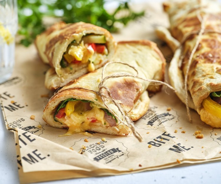 Croque van flatbread met Brugge kaas en geroosterde groenten Een opgerolde croque of tosti van homemade flatbread ligt op een bruin inpakpapiertje. In de openzijde van de croque smelt de kaas mooi uit en zie je een glimp van de geroosterde groenten zoals paprika en courgette.
