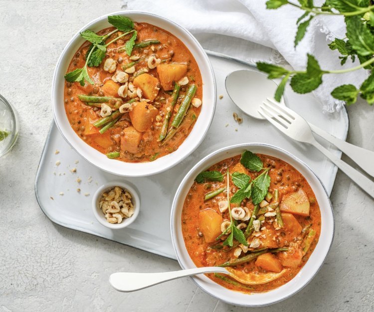 Twee heerlijke curry staan uitgeschept op tafel. Ze hebben een mooie oranje kleur door, jawel, de curry pasta. Ze staan beiden op een poseleinen dienplateau met bestek en een potje pina- en hazelnoten. 