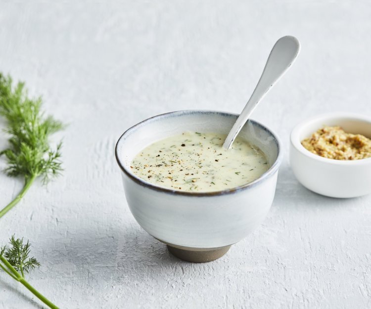 Dillesaus met mosterd De dillesaus is geserveerd in klein porselein potje, er zit een witte lepel in en een takje dille langs.