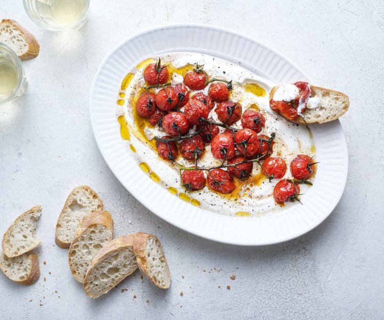 Dip van yoghurt met geroosterde tomaten Een groot ovaal bord is gevuld met de romige yoghurt waarop de dieprode kerstomaatjes bovenop liggen. Er liggen enkele sneetjes stokbrood naast om te dippen. Er staan ook twee glaasjes witte wijn in beeld.
