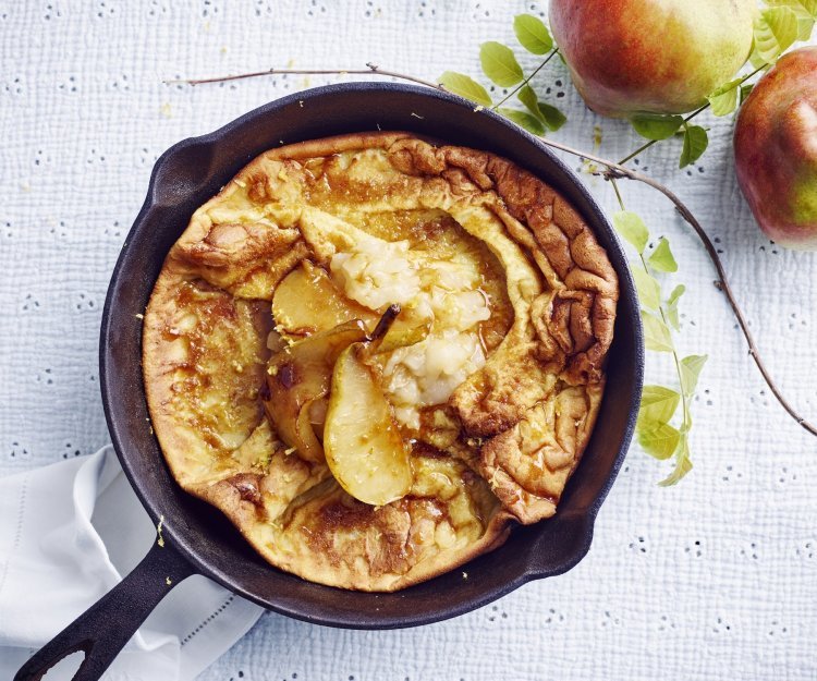 Dutch baby met gekarameliseerde peer en perencompote Een ovenpannenkoek met heerlijke peertjes, hij komt net uit de oven en ligt nog in de pan. Er liggen 2 verse peperen langs.