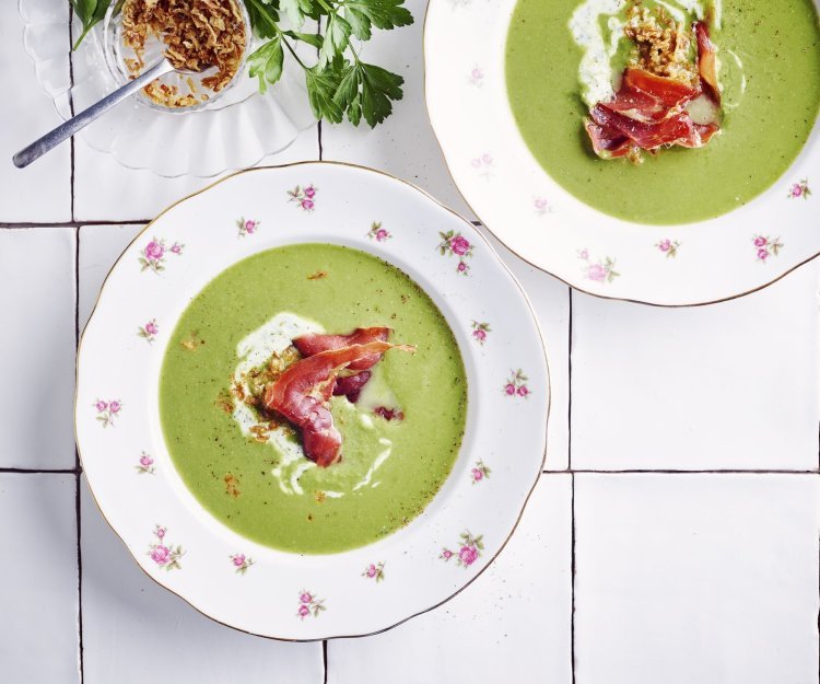 2 porties erwtensoep staan klaar op het werkblad van de keuken. De erwtensoep heeft een mooie groene kleur die mooi contrast geeft in de witte borden. De soep is afgewerkt met peterselieyoghurt, krokante ham en gefruite uitjes.