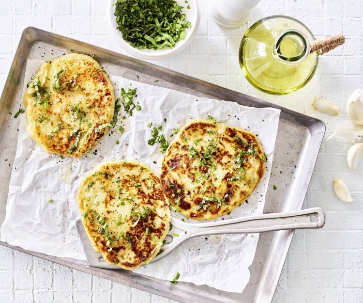 Flatbread van aardappel Drie broodjes zijn gebakken, en wat voor een! Flatbread gemaakt van een restje aardappelpuree liggen te blinken op een grijze bakplaat met wit boterpapiertje op. Verse kruiden liggen klaar om af te werken.