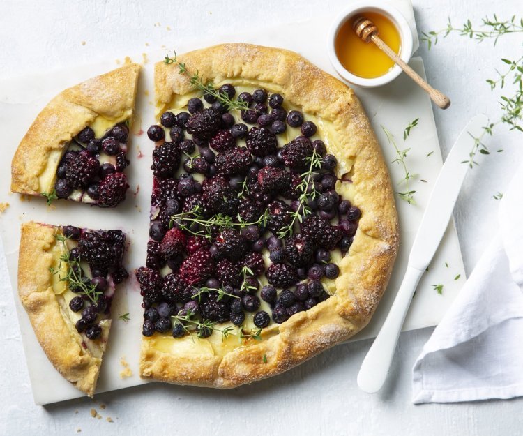 Galette met bramen, blauwe bessen en Belgische brie ligt aanhesneden op het aanrecht. Vergezeld met een potje gouden honing en een tijmplantje. 