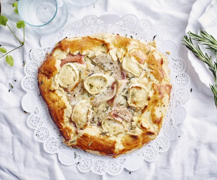 Galette met geitenkaas, peer en ham Een plaattaart of galette ligt op een rond taartpapiertje, klaar om aangesneden te worden. Er liggen enkele takjes rozemarijn langs voor de afwerking.