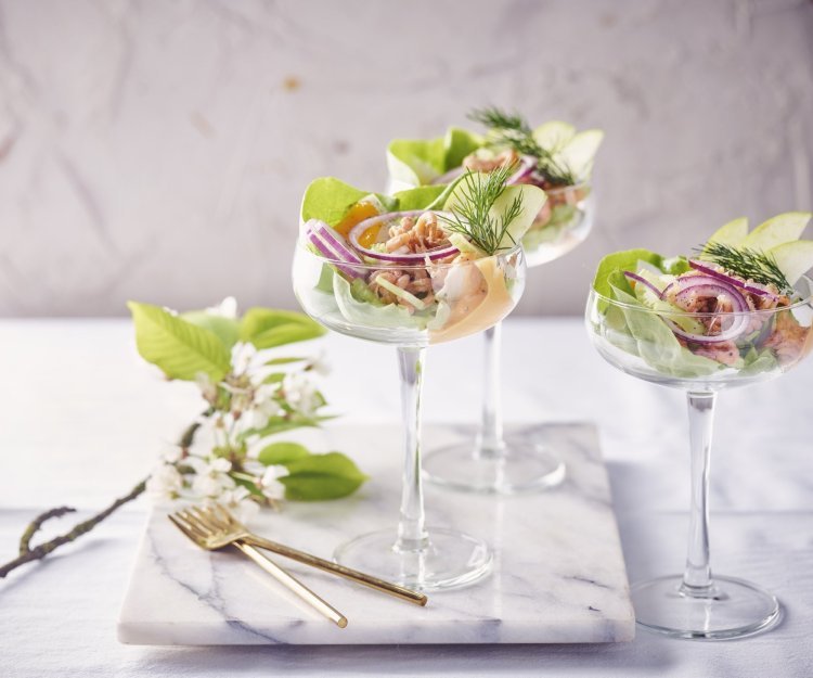 Garnalencocktail met appel en selderij Drie garnalencocktails staan geserveerd op een marmer aanrecht, in mooie wijnglazen met een brede opening bovenaan. Er liggen gouden vorkjes naast om het feestgehalte een extra boost te geven.