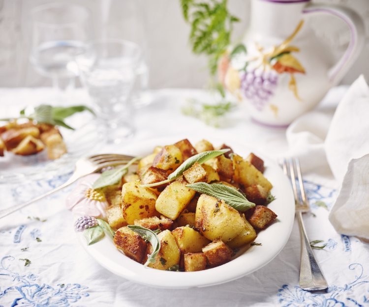 Gebakken aardappelen liggen in een groot wit bord op een gedekte tafel. De verse salieblaadjes geven de krokante aardappelstukjes extra kleur. 