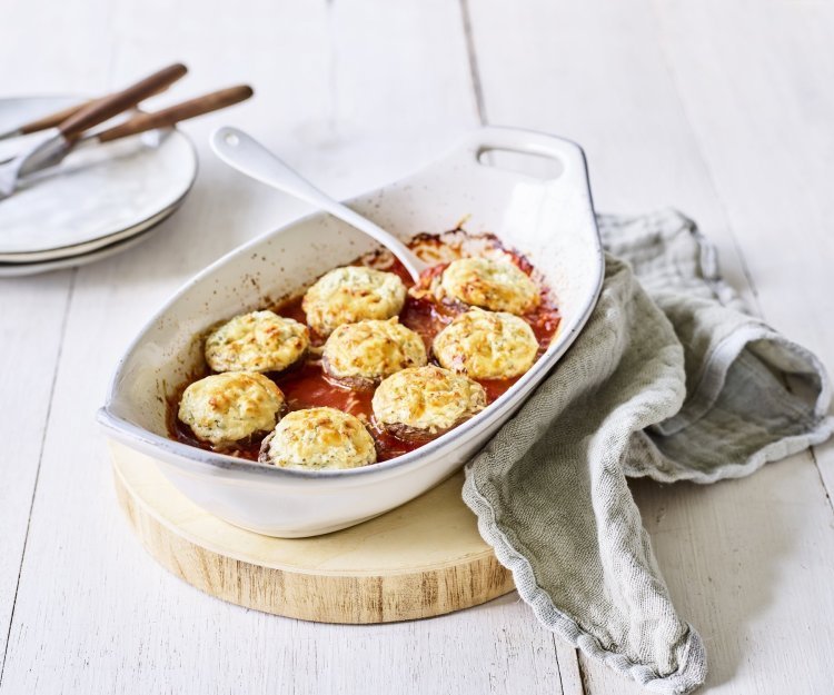 Witte ovenschotel staat op een houten snijplankje op tafel. De ovenschotel is gevuld met de rode tomatensaus waarin de champignons mooi in geschikt zijn. Achteraan de foto zie je bordjes staan met bestek om aan de slag te gaan. 