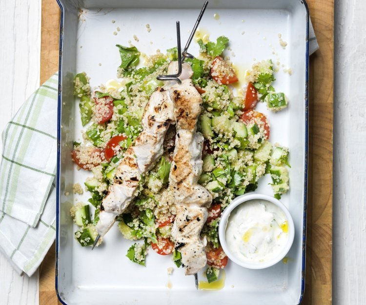 Grote witte bakplaat staat op tafel met eerst een quinoaslaatje en groentjes gepresenteerd.  Hierop liggen de gegrilde brochettes van kip met een dressing van yoghurt. 