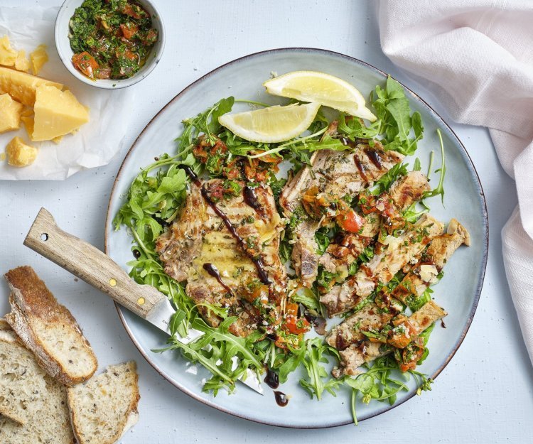 Gegrilde kotelet met tomatengremolata en oude kaas Op een groot blauw/grijs bord ligt eerst een salade van rucola waarop de mooi gegrilde kotelet van kalfsvlees op ligt. De kotelet is in reepjes gesneden waarop de gremolata van tomaten en de oude kaas over gestrooid is. Naast het bord liggen de brokkelkaas en de rest van de gremolata.
