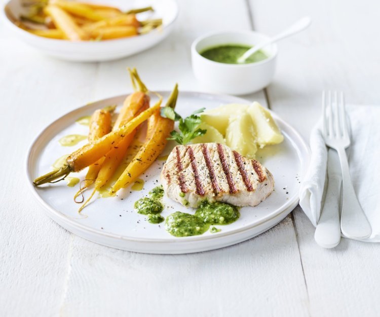 Een mooi in streepjes gegegrild stukje varkensvlees ligt op een wit bord, geserveerd met gegaarde wortels en gekookte aardappelen. We serveren geen saus bij dit stukje vlees, maar homemade pesto van peterselie en Brugge kaas. 