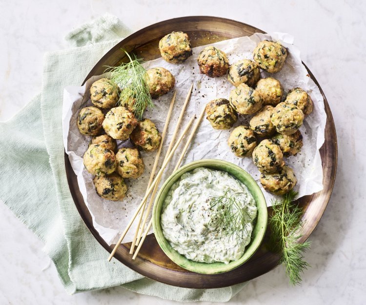 Op het aanrecht staat een houten ronde serveerschaal met een wit boterpapiertje op. Hierop liggen kleine gehaktballetjes met groene stukjes in van de courgette en spinazie. De dip op basis van tzatziki staat ook op de schaal. Het gerecht is afgewerkt met enkele takjes dille. 