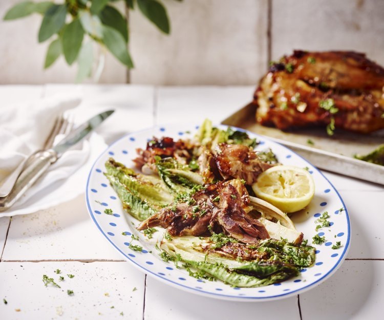 Gelakt hammetje met gegrilde Romeinse salade en gremolata Hammetje ligt op een ovenplaat achteraan in beeld. Op de voorgrond staat een ovaal wit bord met blauwe bloemetjes op de rand. Dit bordje in gevuld met enkele plukken vlees van de gelakte schenkels en gegrilde romeinse sla.