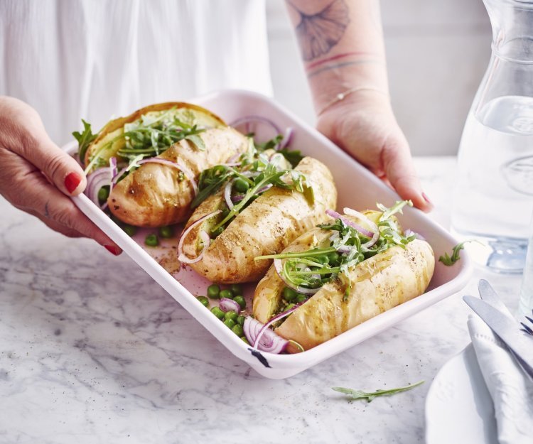 Gepofte aardappel met pesto, erwtjes en rucola 3 gepofte aardappelen met pesto, erwtjes en rucola liggen in een witte ovenschaal. De vrouw wil de ovenschaal plaatsen op tafel.