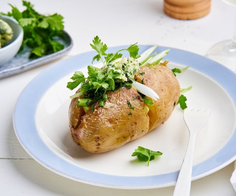 Een gepofte aardappel ligt op een bordje, gevuld met homemade kruidenboter, lente-ui en verse kruiden. 