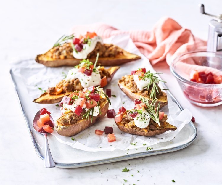 Gepofte zoete aardappel gevuld met gehakt, rode biet en tomaten Op het aanrecht staat een rechthoekige porseleinen serveerschaal met hierop de gevulde zoete aardappelen. Het gerechtje heeft een kleur waar je blij van wordt! Het witte toefje van de zure room, blokjes rode biet en tomaat en afwerken doen we met een frisgroen takje rozemarijn. Een prachtig gerecht in smaak en kleur!