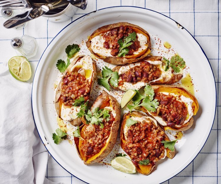 Een ronde ovenschaal met de gepofte zoete aardappelen staat op tafel. De gepofte aardappelen zijn gevuld met yoghurt en hierbovenop de chili sin carne, ze zijn afgewerkt met enkele blaadjes koriander en partjes limoen. De witte ovenschaal heeft een blauw randje en staat op een blauw geruite handdoek.