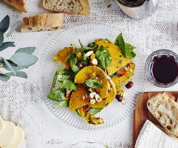 Een glazen bordje staat op tafel met geroosterde partjes pompoen. De salade krijgt een mooi groen kleurtje door de verse spinazie, hij is afgewerkt met enkele hazelnoten. Op en top een herfstsalade boordevol smaak.