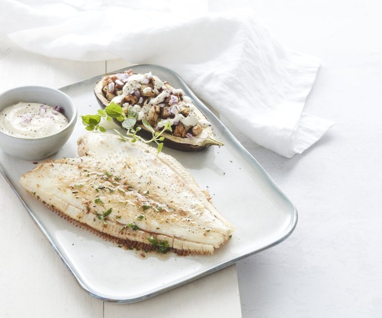 Een gebakken schartong ligt op een rechthoekig wit bord. De halve aubergine ligt naast de gebakken vis, hij is gevuld met stukjes knolselder, peer en walnoten. Het gerechtje is afgewerkt met verse gehakte kruiden en een lepeltje zure room.