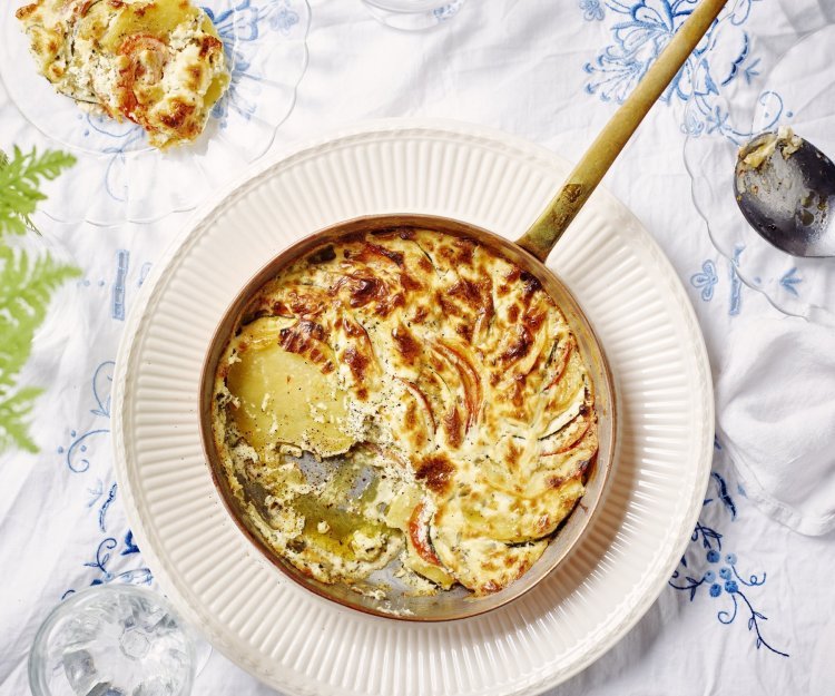 Gratin van aardappelen, courgette, tomaat en kruidenkaas Een heerlijke gratin met groenten en kaas klaargemaakt in een ovenbestendige pan staat in het midden van de tafel. Er is al een portie uitgeschept op een bordje. Op tafel ligt een wit tafelkleed met enkele geborduurde blauwe bloemen.