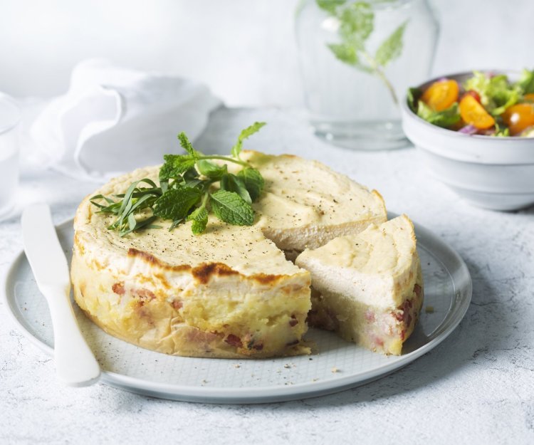 Een hartige cheesecake smaakt heerlijk als lunch of avondmaal. Door het tomatenslaatje krijg je de frisse toets die de cheesesmaak verfrissen. De cheescake ligt aangesneden op een groot rond bord, het slaatje staat langs het bord en een karaf water met takje munt zie je achteraan in de foto. 