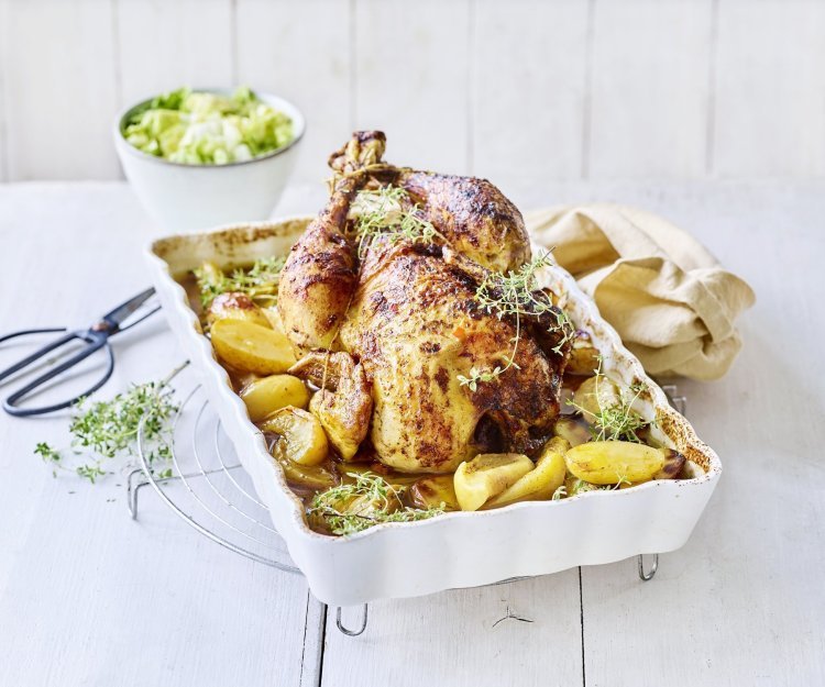 Hoevekip uit de oven met appeltjes en krieltjes De gebraden kip ligt te blinken in een ovenschaal met krieltjes en appeltjes naast. Het gerechtje is afgewerkt met groene verse tijmtakjes, het zwarte schaartje waarmee de kruiden werden afgeknipt ligt er langs.