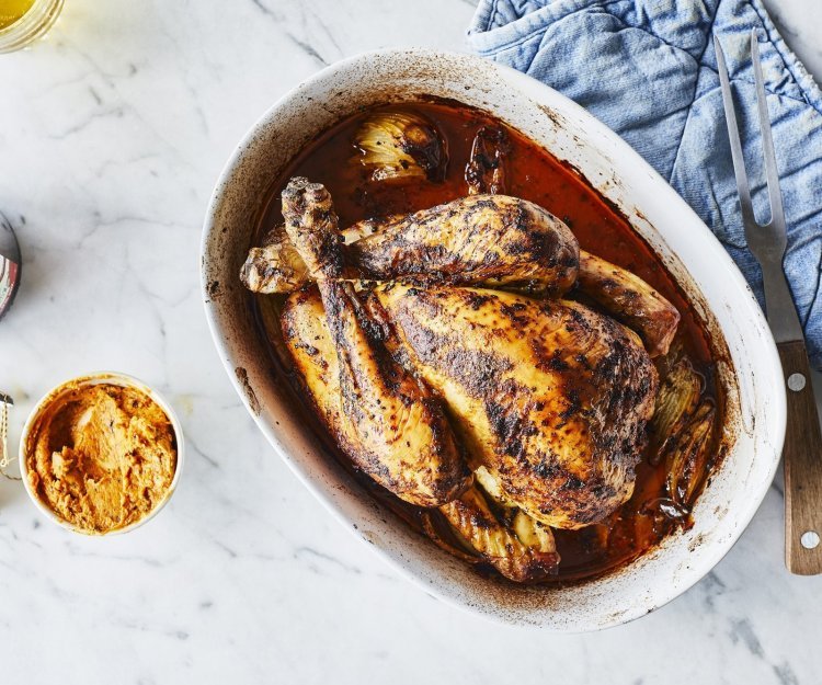 Kip in de oven met cider en beurre café de paris Een heerlijk gegrilde kip uit de oven ligt in een ovenshaal. Ze is heerlijk krokant gebakken en lekker sappig door de homemade kruidenboter. Ze staat op het marmere aanrecht met blauwe nap er langs.