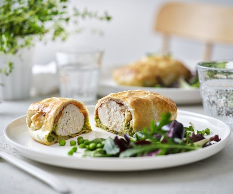 Kip wellington met gedroogde ham en erwtjespuree met munt Een mini wellington van kipfilet ligt opengesneden op een wit bord. Hierin zie je mooi een laagje erwtjespuree onder het witte vlees in het bladerdeeg pakje. Er is nog wat gemengde sla en erwtjes toegevoegd op het bord.