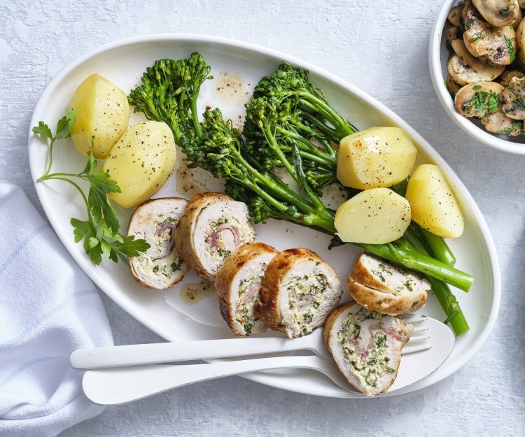 Kipfilet gevuld met een mengsel van roomkaas, mosterd, kruiden en noten. Om af te sluiten voegde we ook nog enkele lapjes spek toe en rolde we hem dicht. Hij ligt aangesneden op een ovaal bord vergezeld van gekookte aardappelen en gekookte broccolini. De gebakken champignons staan in een apart kommetje met een takje peterselie. 