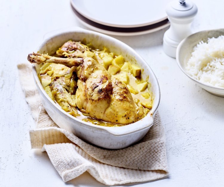 Konijn uit de oven met appel en curry Op tafel staat een witte ovale ovenschotel met hierin de gele currysaus met stukjes appel en ananas. Hier bovenop liggen de bouten te blinken. De ovenschotel staat op een beige linnen handdoek met rijst, borden en een witte pepermolen langs.