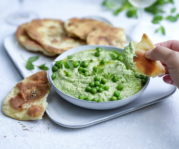 Krokant gebakken platbrood ligt op een serveerschaaltje waarbij een dip van erwten en roomkaas in geserveerd. De dip is gemixt en heeft een mooie groene kleur, om af te werken zijn er nog enkele verse erwtjes bovenop gestrooid. Een hand neem alvast een stukje platbrood om te dippen in gezonde groentedip. 