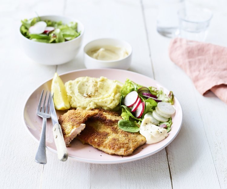 Shnitzel met zelfgemaakte mayonaise Een homemade shnitzel ligt op een pastelroze bord, samen met aardappelpuree, verse sla en zelfgemaakte mayonaise. Er is nog wat extra salade in een wit potje en mayonaise voor extra lekkers op te scheppen.
