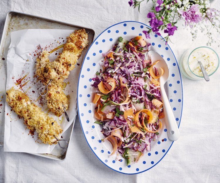 Krokante kipbrochette ligt nog de sidderen op een ovenplaatje. Daarnaast staat een kleurrijk groot bord met een salade op basis van rode kool, wortel en komkommer. De dressing op basis van yoghurt staat langs de salade.