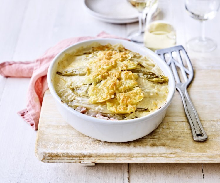 Er staat een ovenschotel op een houten plank, op de tafel. Er ligt een lepel naar de ovenschotel die gevuld is met een lasagne van witloof, ham, kaas en aardappelen. Er ligt ook een coraalroze servette bij.