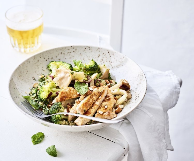 Een rijkgevulde quinoasalade boordevol verse groenten en kruiden zoals broccoli, paddenstoelen, rode ui, peterselie, koriander en munt. Bovenop de quinoa werd het varkensvlees geplaatst in schijfjes. Er staat een witbier bovenaan het bord, op de tafel.