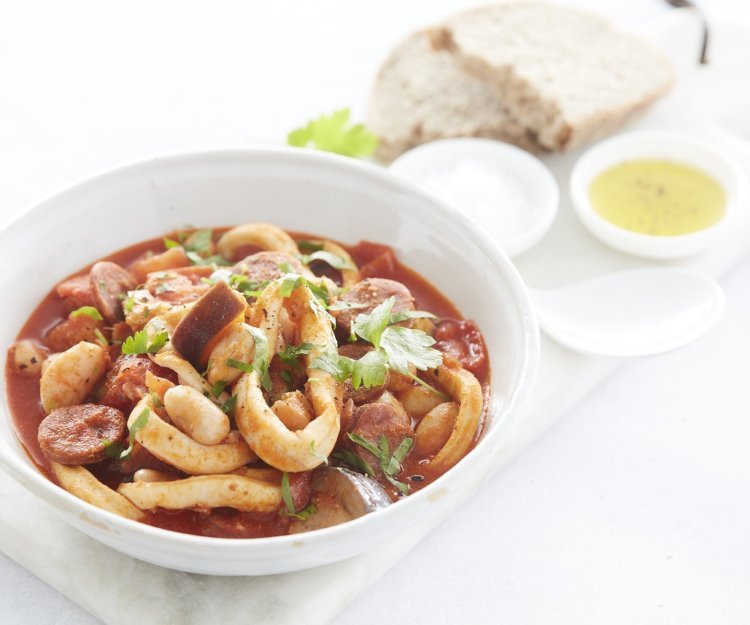 Mediterraans stoofpotje van inktvis en chorizo Een goed gevuld wit bord met het stoofpotje staat klaar om aan te vallen. De kleur is rood van de vele tomaten en chorizo die zijn gebruikt. Het bordje staat op een marmere snijplank waar ook brood en een klein potje olijfolie op staat.