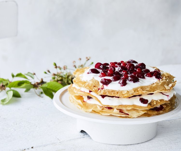 Grote ronde millefeuille met kersen en verse roomkaas. Deze staat op een witte taartschaal op een bleke houten tafel. Op tafel ligt ook een takje groen voor extra sfeer. 