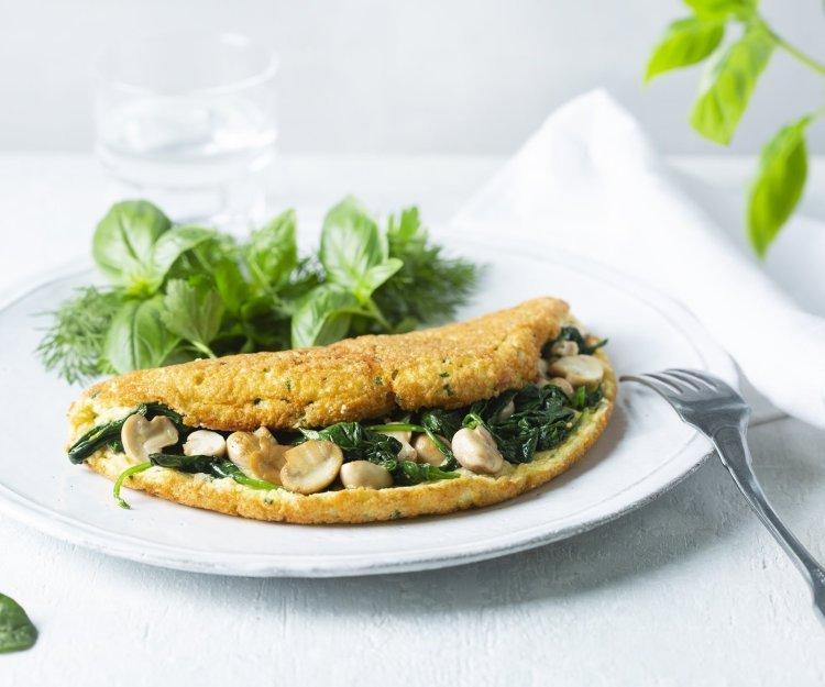 Omeletsoufflé met champignons, spinazie en kaas Een heerlijke dichtgevouwde omeletsoufflé gevuld met champignons, spinazie en kaas ligt op een wit bord. Het gerechtje werd afgewerkt met een slaatje van verse kruiden, denk maar aan basilicum en dille.