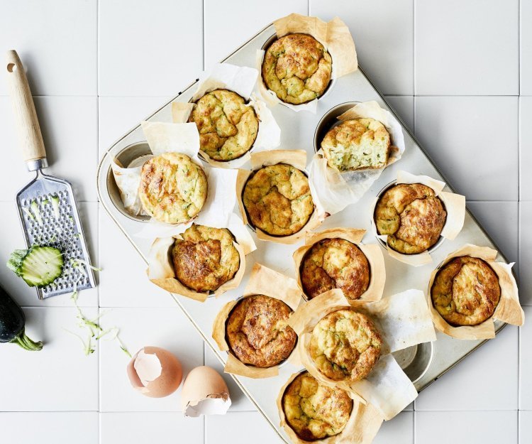 Ontbijtmuffins met courgette, yoghurt en Belgische kaas in een grote muffinvorm. Klaar om eruti te halen en op te eten. Naast de bakvorm ligt een verse courgette en lege eierschalen voor de sfeer.