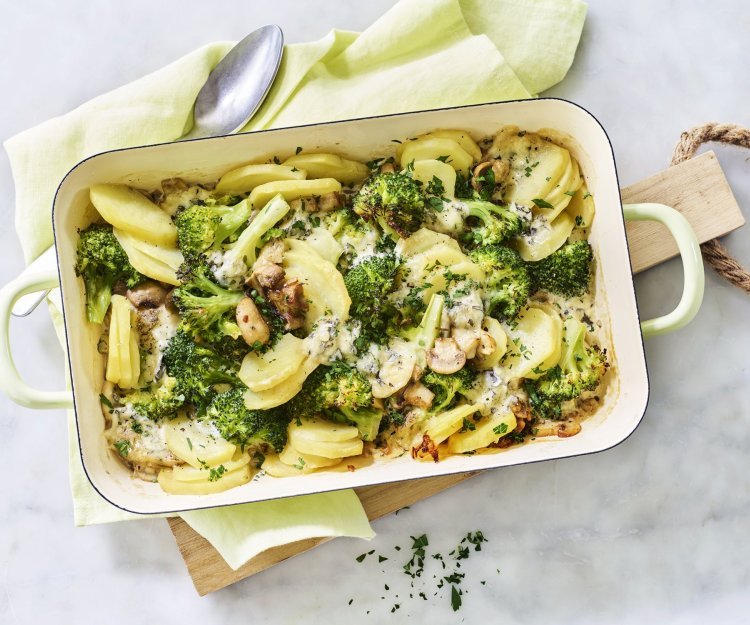 Ovenschotel van aardappel, broccoli, champignons en blauwe kaas Een ovenschotel van aardappel, broccoli, champignons en blauwe kaas staat op het midden van het marmere aanrecht op een houten snijplank. Verse kruiden zorgen voor een extra groene touch!