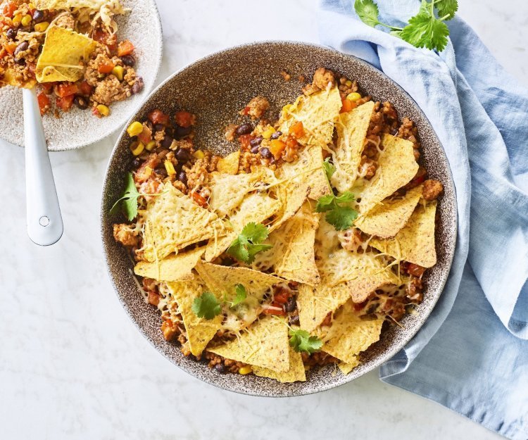 Ovenschotel van chili con carne met nachos en kaas Onderaan de ronde ovenschaal ligt de chili con carne met bovenop de nachos. Er is al een portie uitgeschept op een wit bordje en staat langs de ovenschaal. Er ligt ook een blauwe servette en enkele blaadjes kroiander voor de kleur.