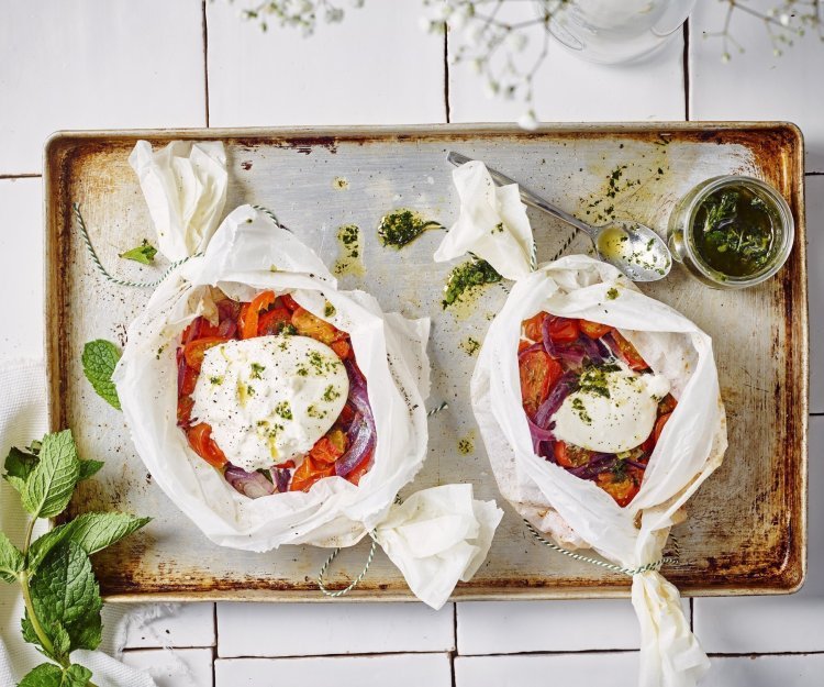 Twee papillotten liggen op een ovenplaat. De papillot is gemaakt in een wit boterpapiertje waarin de groenten een mooi contrast in geven. Denk maar aan de rode paprika's en tomaten. De burrata ligt er bovenop en de muntolie staat in een bokaaltje naast de papillotten.