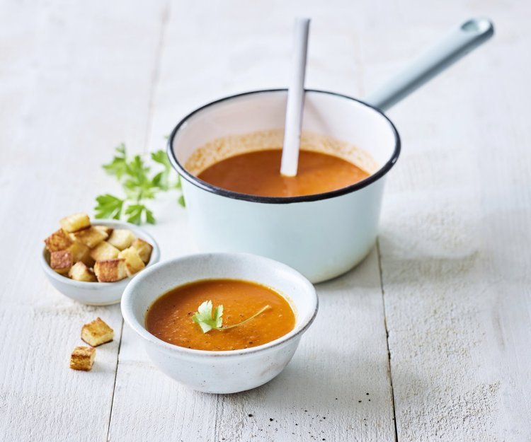 Op een houten witte tafel staat een kopje paprikasoep. De soepkom staat ook in beeld, dit is een lichtblauwe pot die mooi contrast geeft met de oranje-rode soep. De soep is afgewerkt met verse peterselie en croutons.